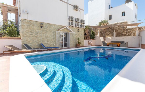 Une piscine aux carreaux bleus, un patio couvert avec une table et des chaises, et un bâtiment équipé de climatiseurs.