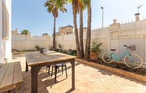 Une terrasse avec une table et des chaises en bois, un vélo turquoise et une clôture blanche agrémentée d’éléments décoratifs.