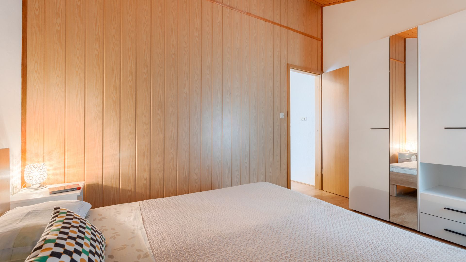 A bedroom with a wooden panel wall, a bed with a patterned pillow, a bedside table with a lamp, and a wardrobe with a mirror.