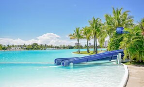 Zona de piscina con tobogán de agua, palmeras y un cielo azul despejado.