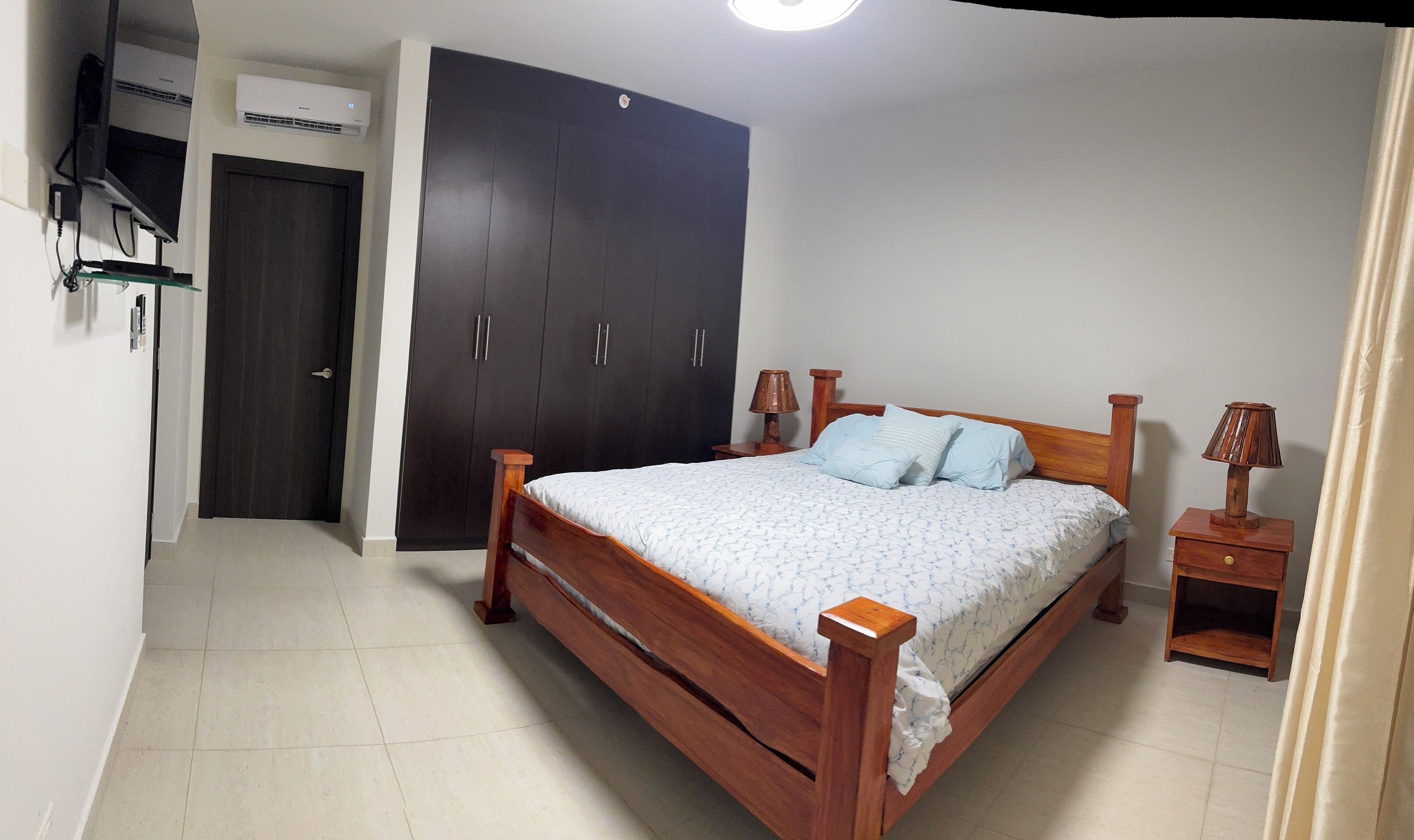 A bedroom with a wooden bed, two bedside tables with lamps, a wardrobe, and an air conditioning unit.