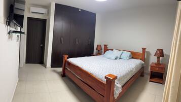 A bedroom with a wooden bed, two bedside tables with lamps, a wardrobe, and an air conditioning unit.