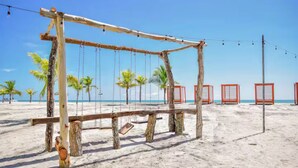 A wooden swing set on a sandy beach with palm trees and a clear blue sky in the background.