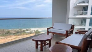 A balcony with a view of the ocean, wooden furniture, and a white cushioned chair.