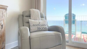 A reclining chair with a 'welcome home' pillow, a wooden cabinet, and a balcony with a railing.