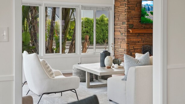 A modern living room with white furniture, a stone fireplace, and large windows overlooking greenery.
