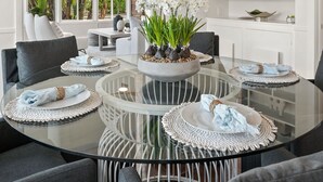 A modern dining area with a glass table surrounded by grey upholstered chairs, a crystal chandelier, and a potted orchid centerpiece.