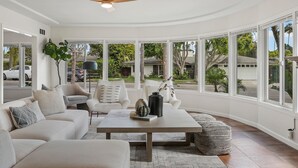 A modern living room with a white sofa, armchairs, a coffee table, and large windows offering a view of the neighborhood.