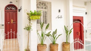Un edificio blanco con puertas rojas y un pequeño ventanal con diseño de celosía. Hay plantas en macetas frente al edificio.