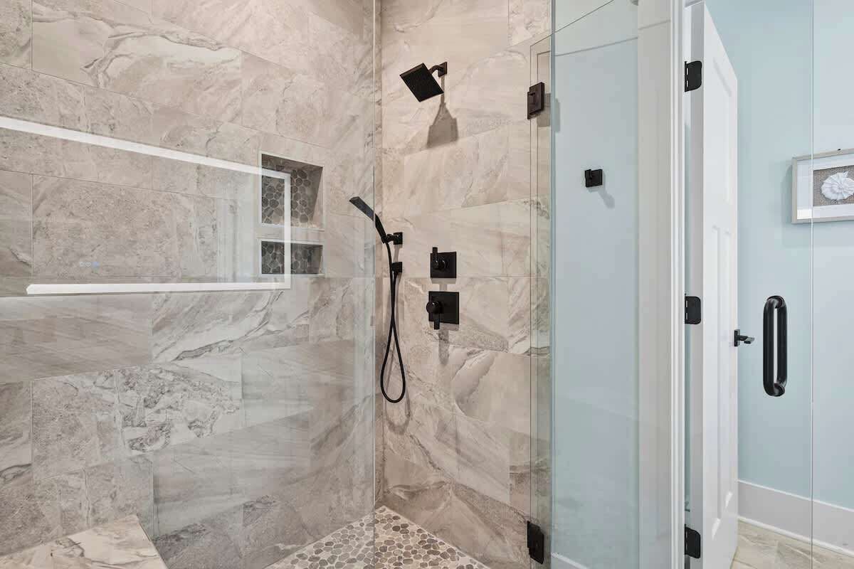 Ducha con cerramiento de vidrio, pared de mármol, un estante pequeño y una puerta con manija.