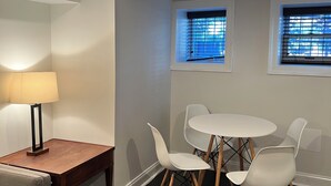 A modern dining area with a wooden table and white chairs, a lamp, and two windows with blinds. - Charming, modern 1-bedroom English Basement in heart of DC with WiFi, AC (Washington)