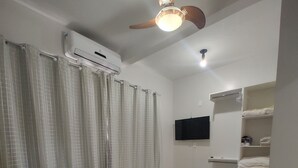 A bedroom with a bed, a ceiling fan, an air conditioning unit, a television, and a shelf with folded clothes.