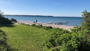 A beach with a wooden pier extending into the water, a grassy area in the foreground, and a clear blue sky. - Amazing apartment in Asperup with sauna (Asperup)