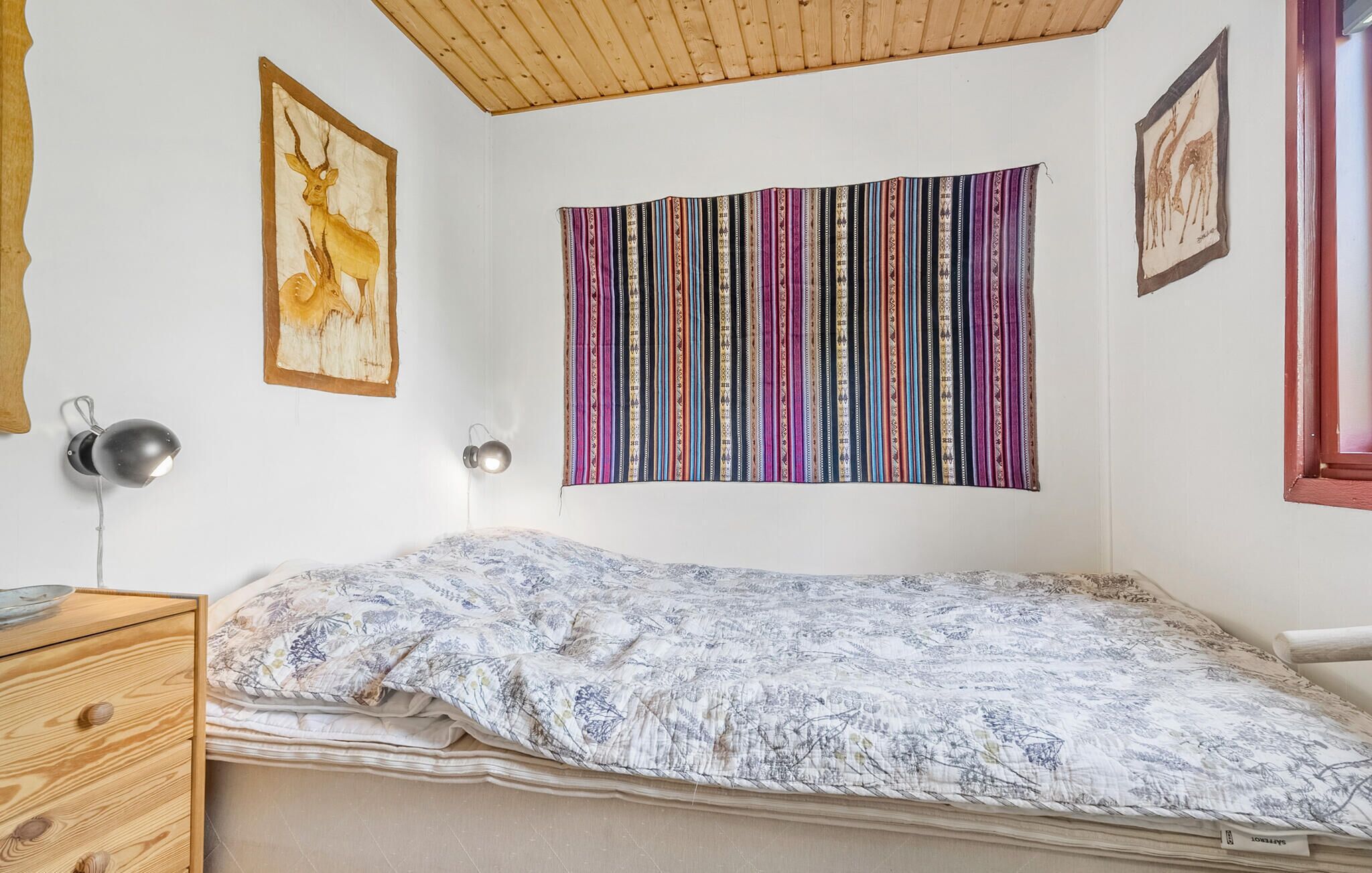 A bedroom with a bed, a wooden dresser, a wall-mounted lamp, and a colorful striped curtain.