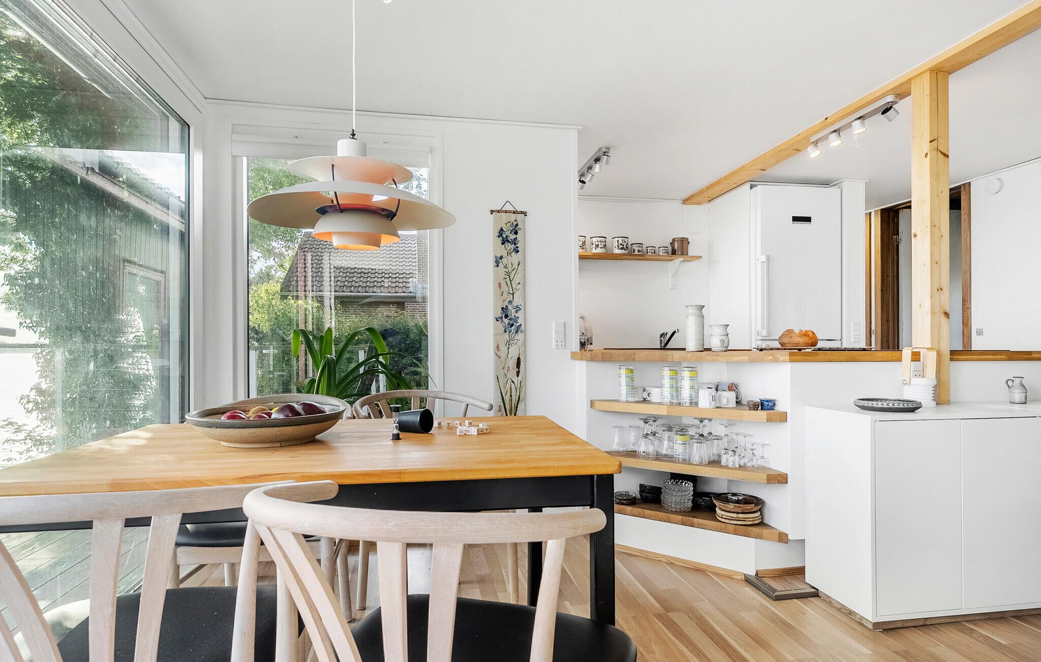 A modern kitchen with a wooden table, chairs, and a hanging light fixture. There are shelves with kitchenware and a window with a view of greenery.