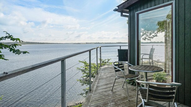 Eine Holzterrasse mit Metallgeländern und Blick auf ein Gewässer. Auf der Terrasse befinden sich zwei Metallstühle und ein kleiner Tisch.