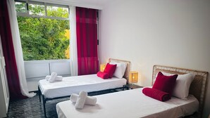 A hotel room with two beds, each with white linens and red pillows, a window with red curtains, and a view of greenery outside. - Cozy 2-Bed & Sofa-bed Studio in Los Cristianos (Playa de la Américas)