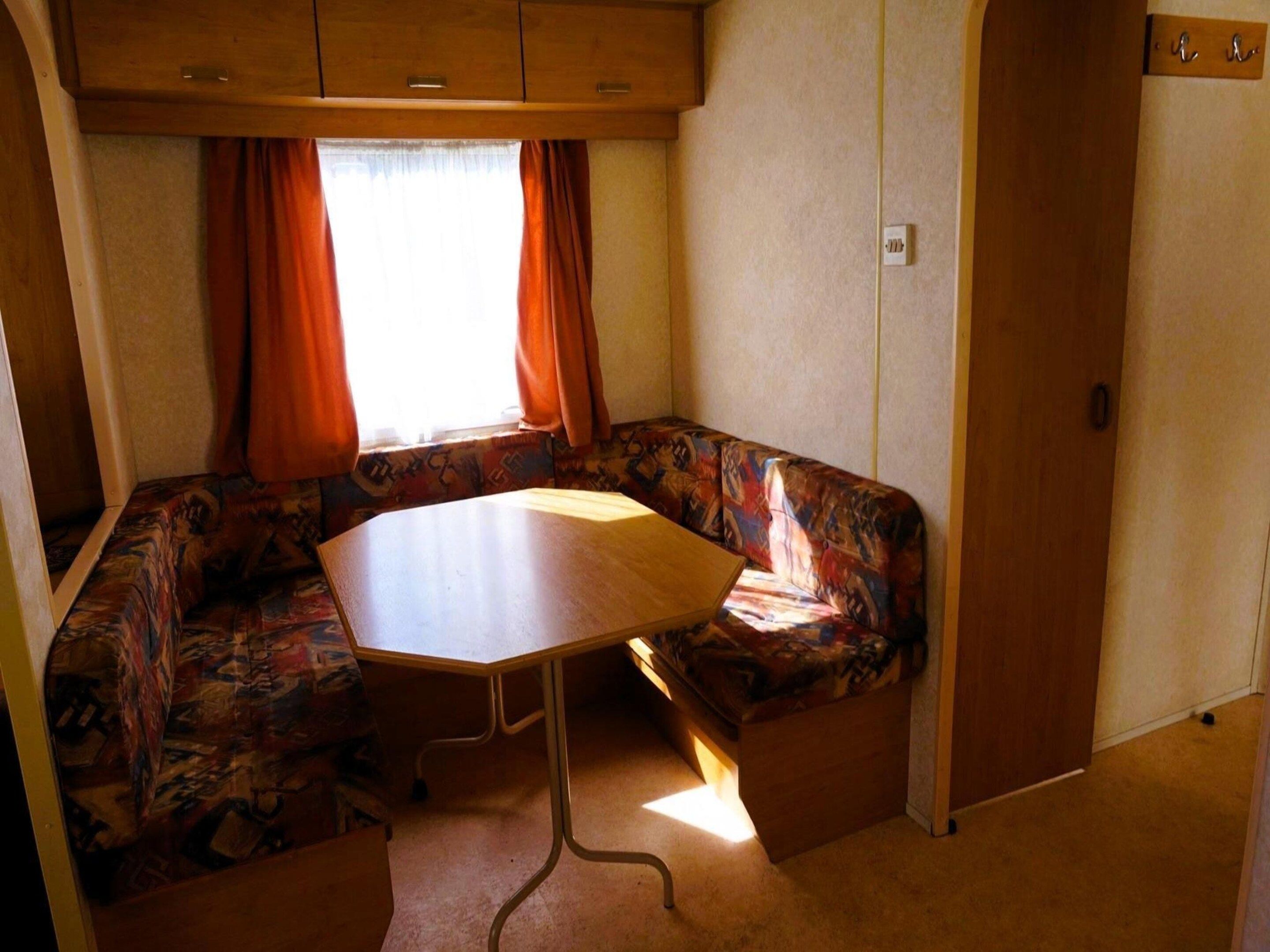 A compact RV interior with a dining table, a cushioned bench, and a window with curtains.