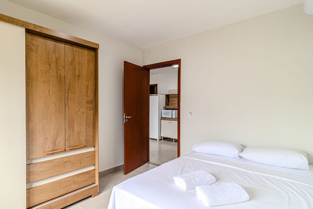 Ein Schlafzimmer mit einem Bett, weißer Bettwäsche, einem Holzschrank und einem durchgehenden offenen Tür erkennbaren Küchenbereich.