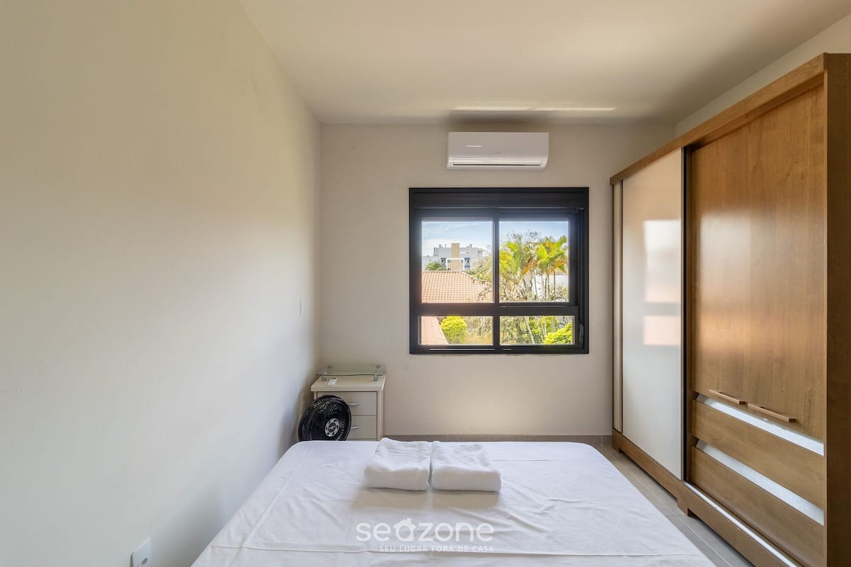 Ein Zimmer mit einem Bett, einem Fenster, einem Holzschrank und einem kleinen Tisch mit einem Ventilator.