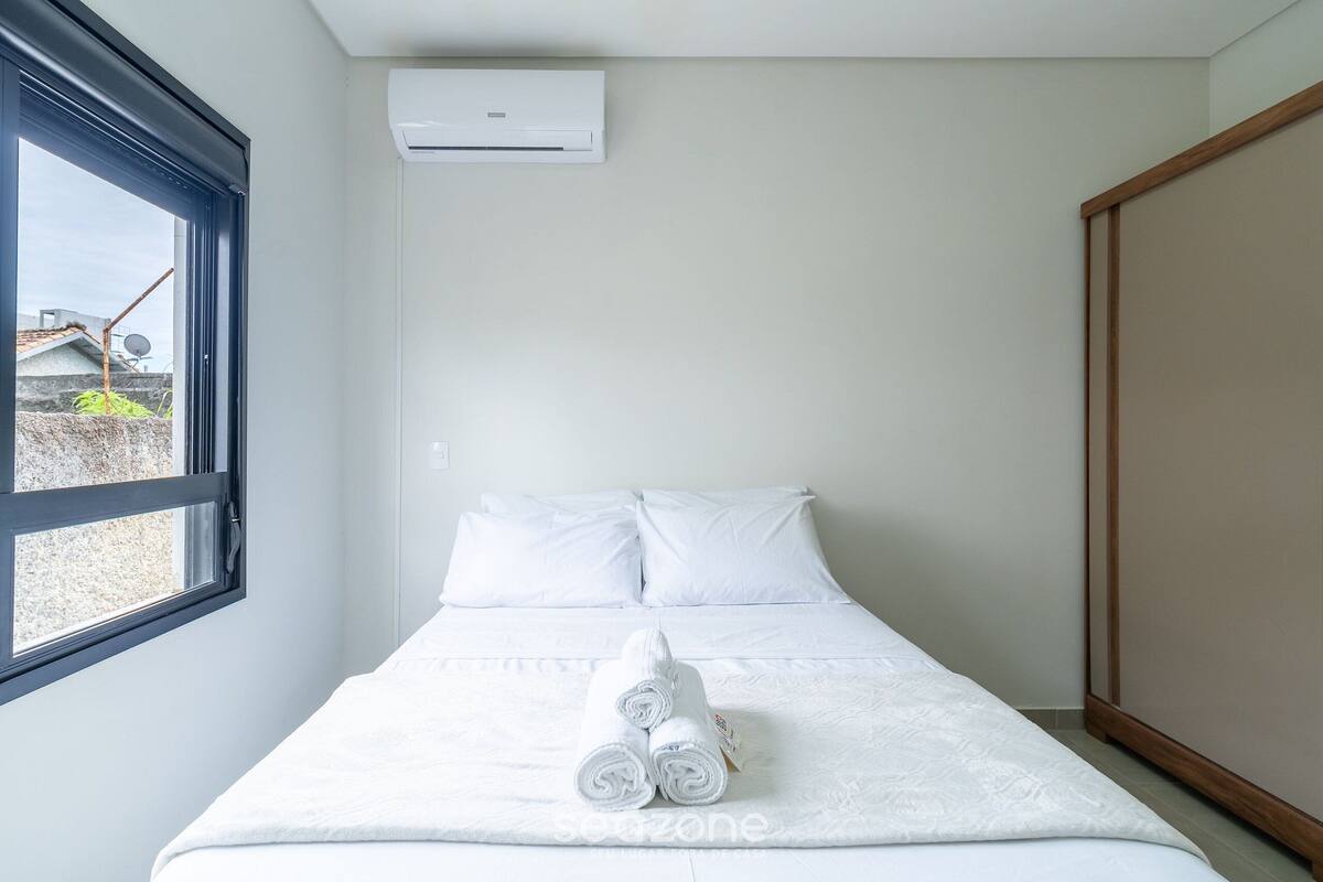 Ein ordentlich bezogenes Bett mit weißen Leintüchern und aufgerollten Handtüchern, eine Klimaanlage an der Wand und ein Fenster mit Blick auf Gebäude.