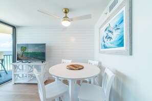 A dining area with a round table, white chairs, a ceiling fan, and a TV displaying 'Emerald Beach Properties'.