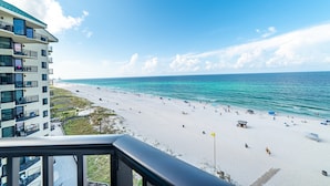 A beach view from a balcony with a clear sky, a sandy beach, and a building with multiple balconies.