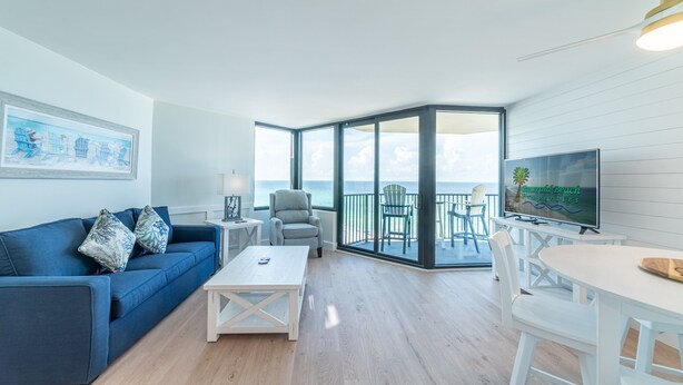 A modern living room with a blue sofa, a white coffee table, and a TV on a white stand. There is a balcony with a view of the sea.