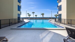 A swimming pool with a clear view of the ocean, surrounded by palm trees and a building with balconies.