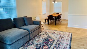 Una sala de estar con un sofá gris, una mesa de comedor de madera con sillas y una alfombra estampada.