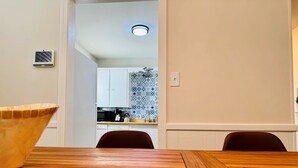 Comedor con una mesa y sillas de madera, cocina al fondo con baldosas estampadas y una lámpara en el techo.