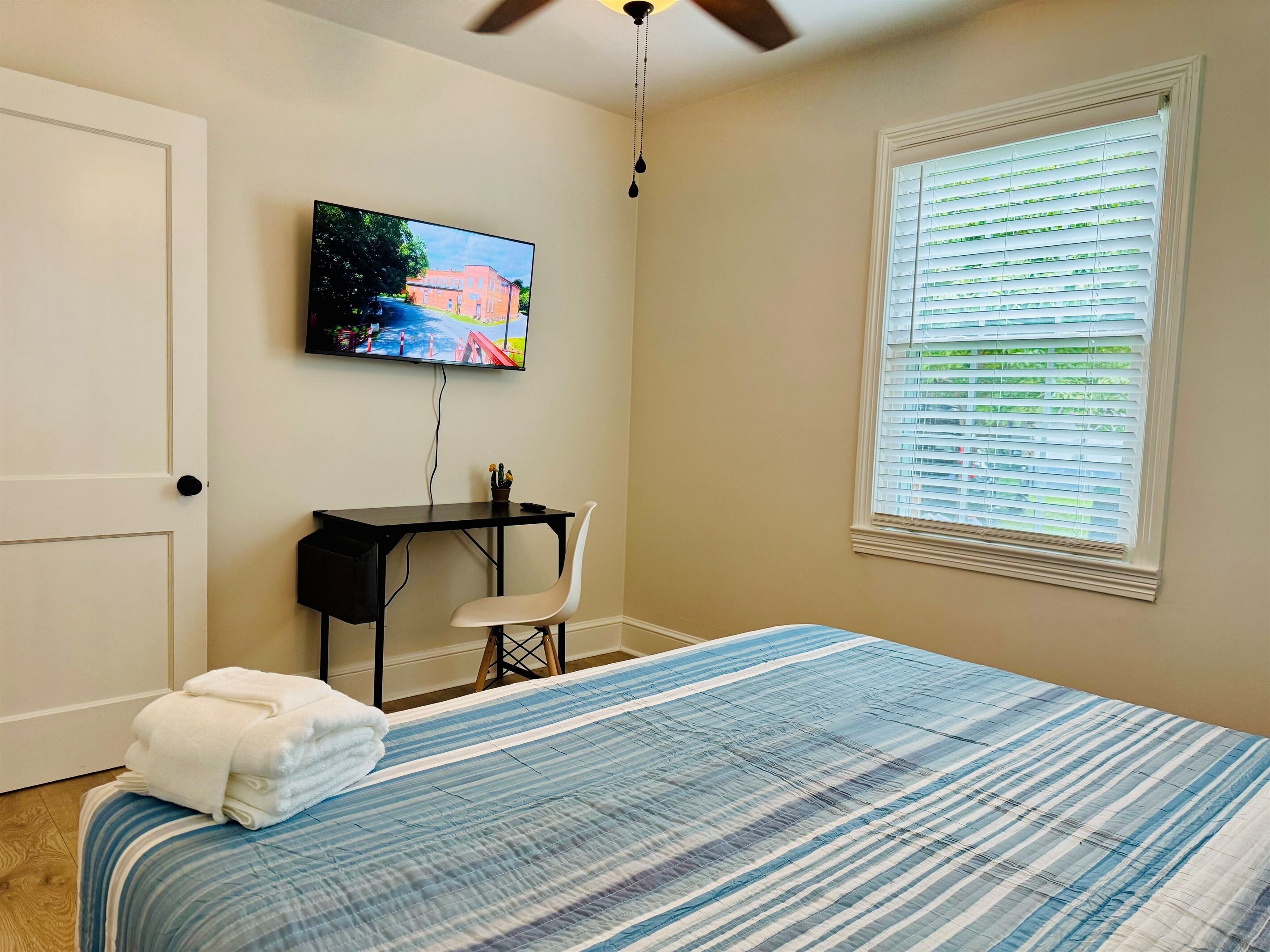 A bedroom with a bed, a desk, a chair, a TV, and a window with blinds.