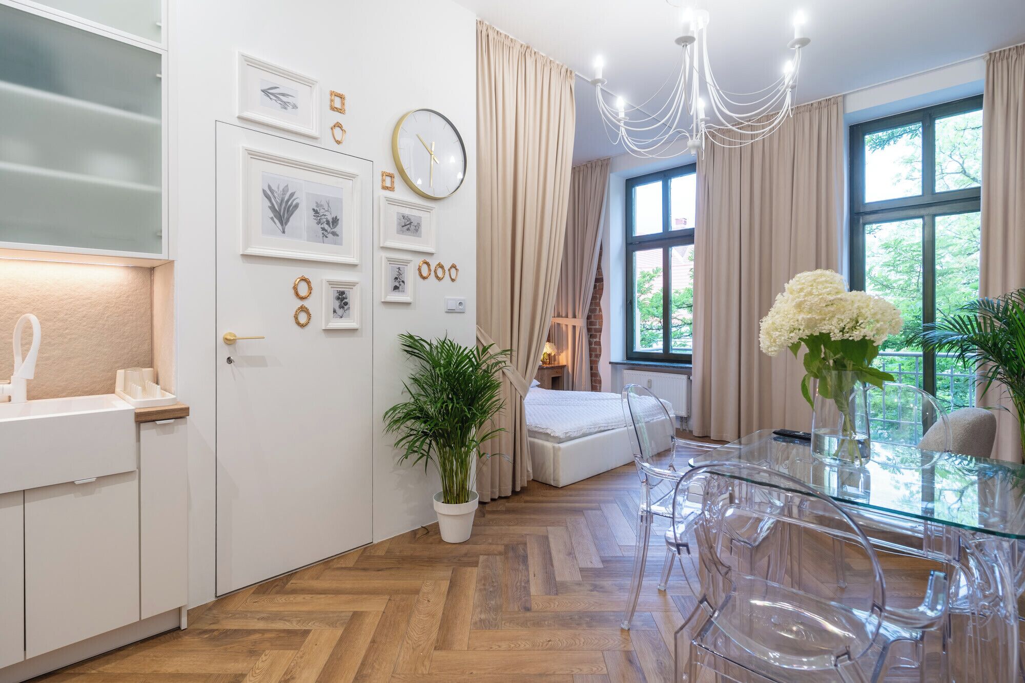 Una habitación de hotel moderna con piso de madera en diseño espiga, una mesa de comedor de vidrio, un sofá blanco y un ventanal con cortinas.