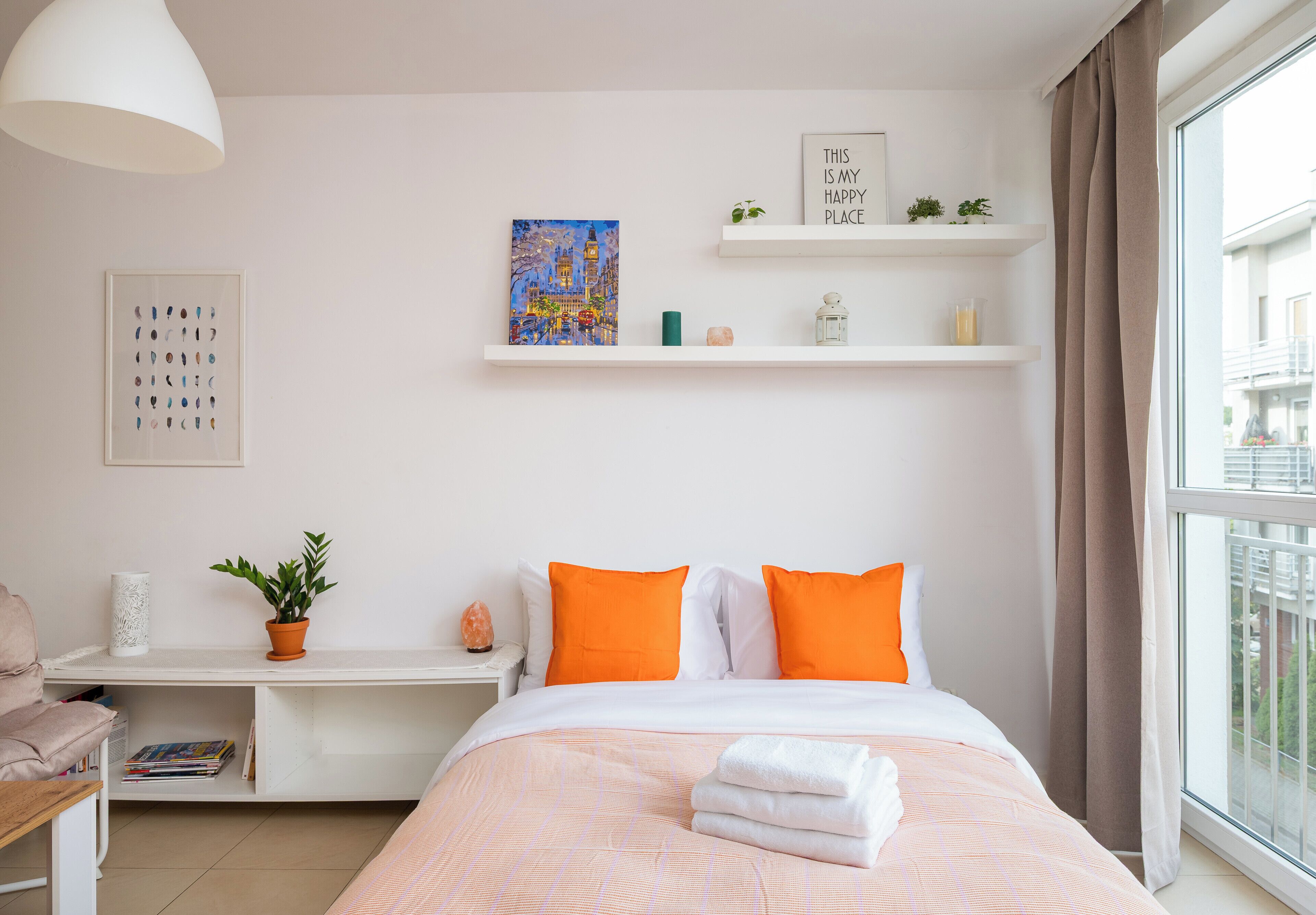 Une chambre à coucher bien rangée, avec un lit, des oreillers, une table de chevet agrémentée d’une plante, une œuvre d’art murale et une étagère avec des objets décoratifs.