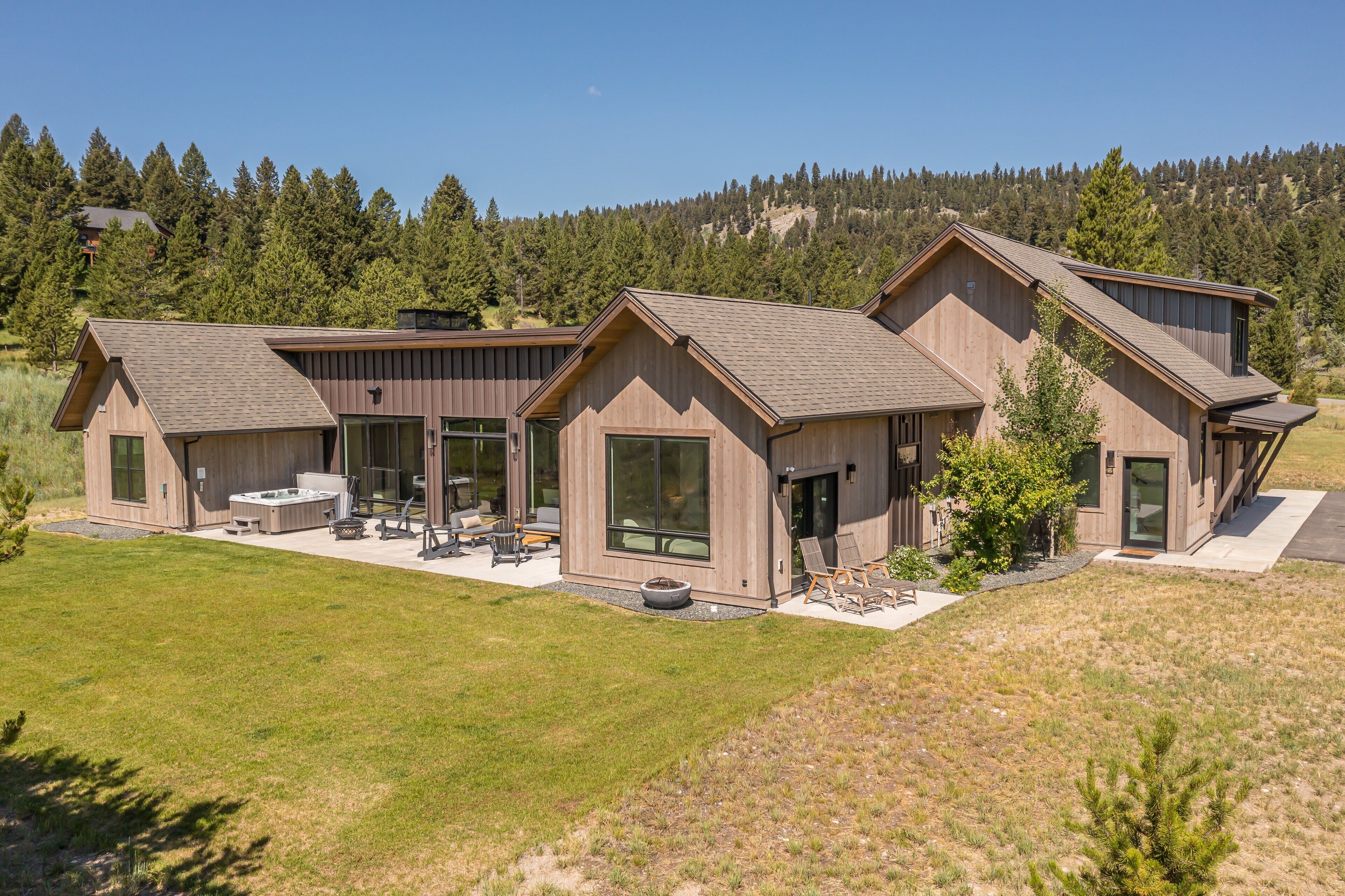 A modern two-story house with large windows, a patio with outdoor furniture, and a view of a forested hillside.