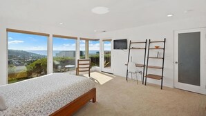 A bedroom with a bed, a desk, and a chair. There are large windows offering a view of the outside.