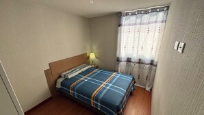 A bedroom with a wooden floor, a bed with a blue and orange striped bedspread, a wooden headboard, a nightstand with a lamp, and a window with a patterned curtain. - In condominium with beautiful views (José Luis Bustamante y Rivero)