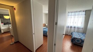 A hotel room with a wooden floor, a bed with a patterned bedspread, a bedside table, and a lamp. There is a door leading to another room. - In condominium with beautiful views (José Luis Bustamante y Rivero)