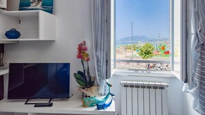 A room with a TV on a white stand, a plant, and a window with a view of a harbor and mountains. - 1 bedroom awesome apartment in Sestri Levante (Sestri Levante)