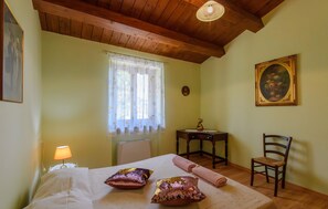 A bedroom with a bed, pillows, a chair, a table with a lamp, a window with curtains, and a painting on the wall.