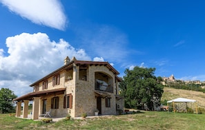 A stone house with a balcony, a covered porch, and a view of a hill with buildings.