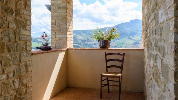 En balkon med stenvægge, en træstol, en potteplante og udsigt til et bjerglandskab.