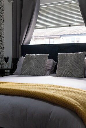 Un lit bien fait, recouvert d’une couverture jaune, avec deux oreillers à motifs chevrons, et une fenêtre avec des stores.