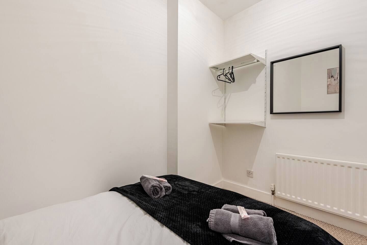 A bedroom with a bed, a black throw blanket, and a towel on the floor. There is a white radiator and a wall-mounted shelf with a mirror and a picture frame.