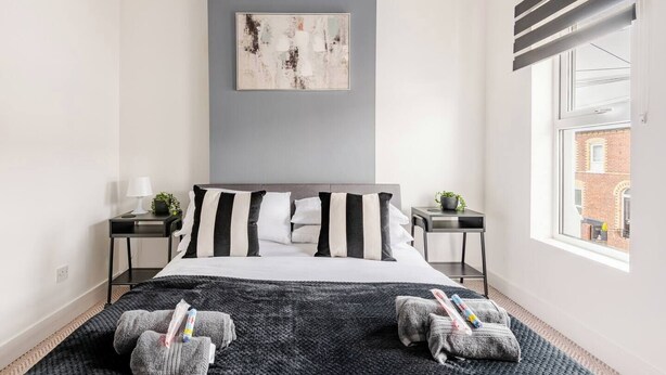A modern bedroom with a large bed, a gray accent wall, a window with blinds, and a view of a building outside. - Modern 2-bed home with free on-street parking, fast Wi-Fi & family amenities. (Belfast)