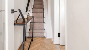 A hallway with a herringbone-patterned wooden floor, a staircase with patterned carpet, and a small table with a glass and a candle. - Modern 2-bed home with free on-street parking, fast Wi-Fi & family amenities. (Belfast)