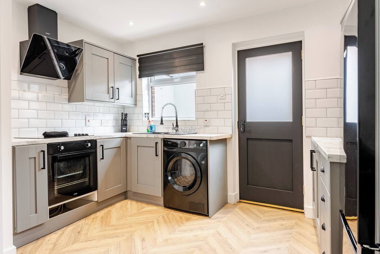 A compact kitchen with a built-in oven, a washing machine, and a window with a black roller blind.