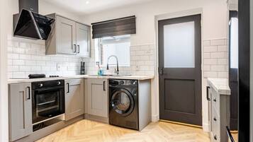 A compact kitchen with a built-in oven, a washing machine, and a window with a black roller blind.