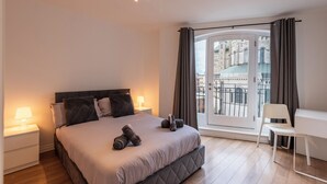 A modern bedroom with a large bed, bedside lamps, a view of a cityscape through a glass door, and a white desk.