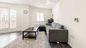 A modern living room with a grey sofa, a black coffee table, and a small plant. There is a clock on the wall and a radiator below a window. - Modern 1-bed apartment in Belfast’s Cathedral Quarter. (Belfast)